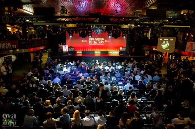 Eine große Menschenmenge, die auf Stühlen vor einer Bühne mit Bildschirmen und Plakaten sitzt, von Scheinwerfern beleuchtet und von Absperrungen umgeben, voller Erwartung auf den Beginn eines Online-Marketing-Rockstars-Events, mit einem Wasserzeichen in der linken unteren Ecke.