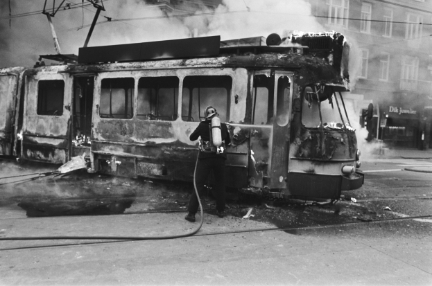 Schwarzes und wei├čes Bild eines Mannes in einem Helm, der neben einer ausgebrannten Stra├čenbahn steht und eine Pfeife h├Ąlt, mit aufsteigendem Rauch und einem Gebäude im Hintergrund.