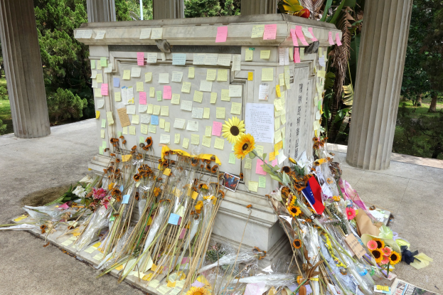 Gedenkstätte mit einem zentralen Pfeiler, der mit Post-its und Blumen geschmückt ist, umgeben von Bäumen, Pflanzen und Gras in einer friedlichen Umgebung.