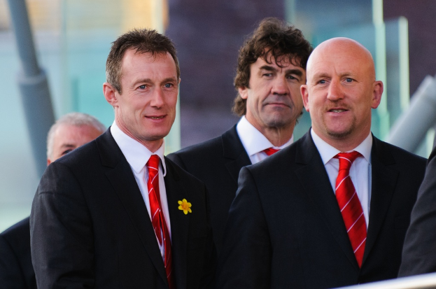 Vier Männer in Anzügen stehen im Vordergrund, mit einem unklaren Hintergrund und einem Pfahl links.