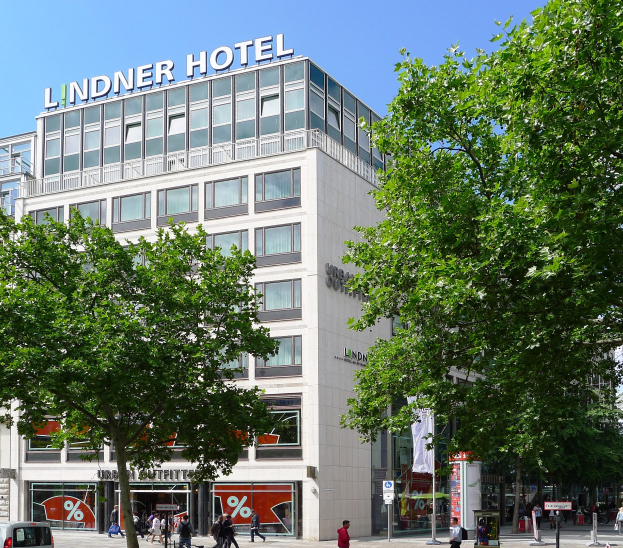 Großes modernes Hotelgebäude mit Glasfenstern und Aufschrift auf der Oberseite, umgeben von Bäumen und einer belebten Straße mit Fußgängern und Fahrzeugen unter einem sichtbaren Himmel.