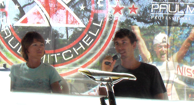 Eine Gruppe von Menschen auf Stühlen, ein Mann mit einem Mikrofon, ein Banner mit einer Person in einer Mütze und ein Fahrradsitz im Vordergrund.