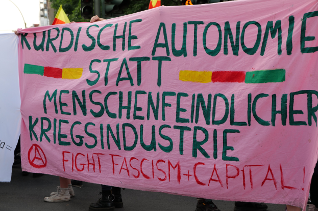 Eine Gruppe von Menschen hält eine pinkfarbene Fahne mit der Aufschrift "Kurdische Autonomie Statt Menschenfeindlicher Kriegsindustrie Fight Fascism Capital" auf einer Straße mit Verkehrszeichen, Fahnen, Bäumen, Gebäuden und einem klaren blauen Himmel im Hintergrund.