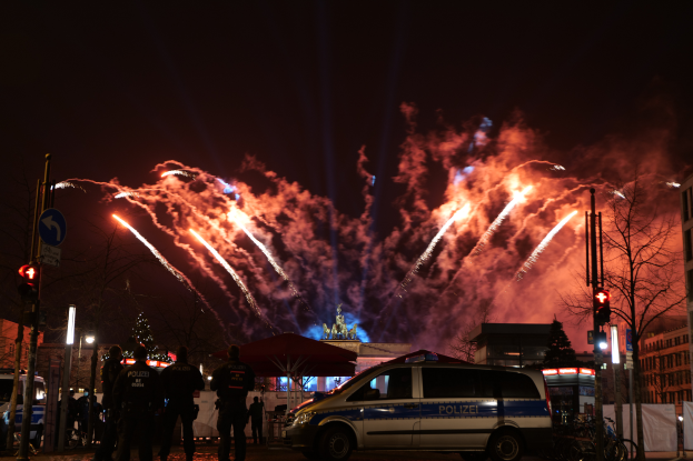 Ein Polizeiwagen steht vor einer Menschenmenge, mit beleuchteten Gebäuden, Bäumen, Polen, Verkehrszeichen, Schildern und Zelten im Hintergrund, unter einem Himmel voller farbiger Feuerwerke.