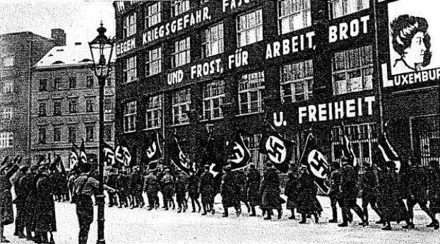 Schwarz-weißes Bild einer Gruppe, die mit Fahnen und Transparenten die Stra├če entlangmarschiert, mit einem Lichtmast links und einer Tafel rechts mit der Aufschrift "Deutsche Nationalsozialistische Arbeiterpartei in Berlin".