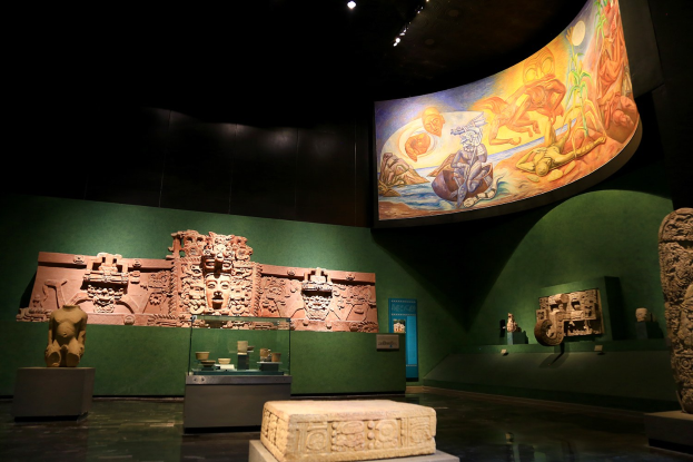 Ein Museumsraum mit verschiedenen Artefakten, darunter Skulpturen, Tischgegenstände, eine Texttafel, eine Wandmalerei und Deckenleuchten im Anthropologischen Museum von Mexiko-Stadt.