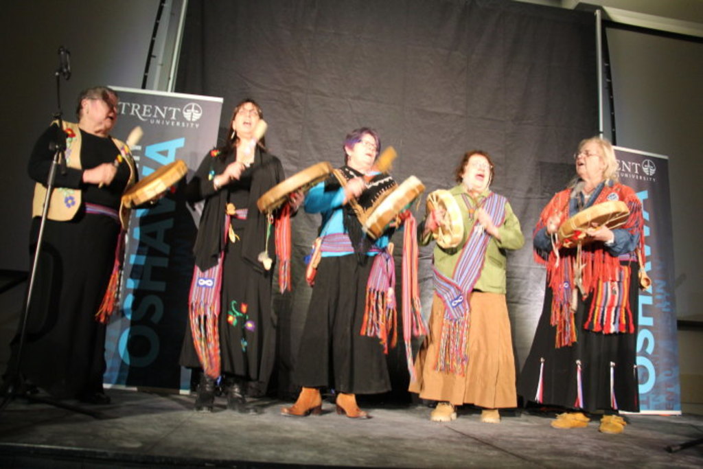 Eine Gruppe von Frauen in bunten Kleidern, die singen und Trommeln halten, mit einem schwarzen Vorhang und zwei blauen Rollplakaten im Hintergrund.