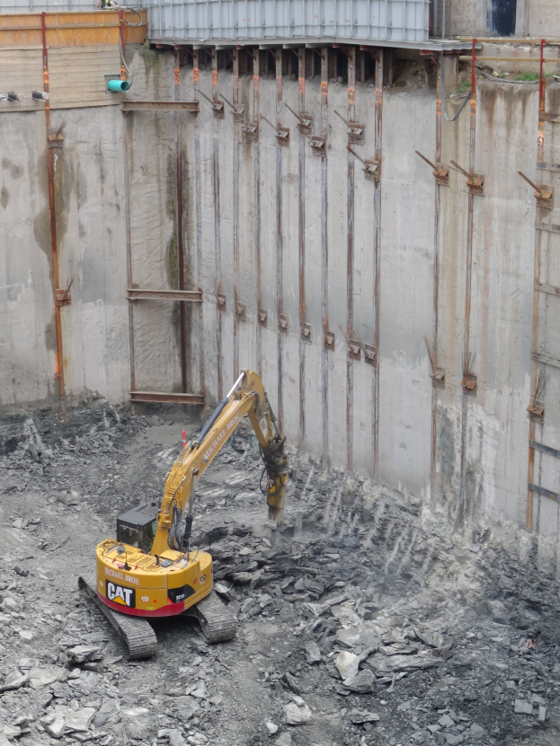 Bagger bei einer Baustelle mit Steinen und Eisenstangen, mit einem teilweise errichteten Gebäude links im Bild.