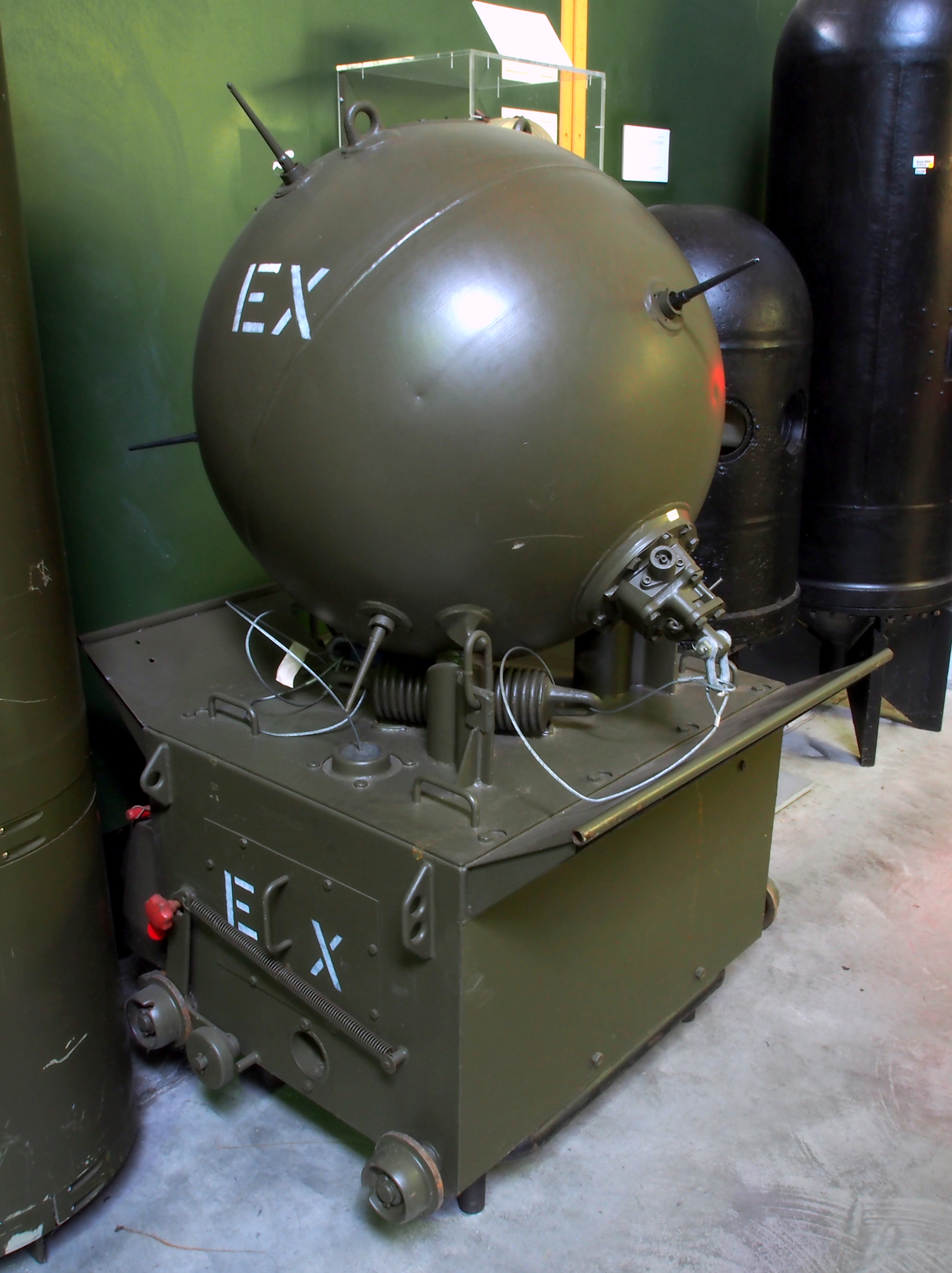 Eine große grüne Splitterbombe mit zylindrischem Metallkörper steht auf einem Tisch umgeben von anderen Gegenständen, mit einer Wand im Hintergrund und Text am unteren Bildrand.