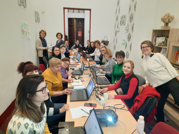 Eine Gruppe von Frauen in einem Workshop für Menschen mit Behinderungen, die um einen Tisch mit Laptops, Papieren und anderen Gegenständen sitzen, mit Jacken auf Stühlen, einem Bücherregal und Papieren an der Wand.