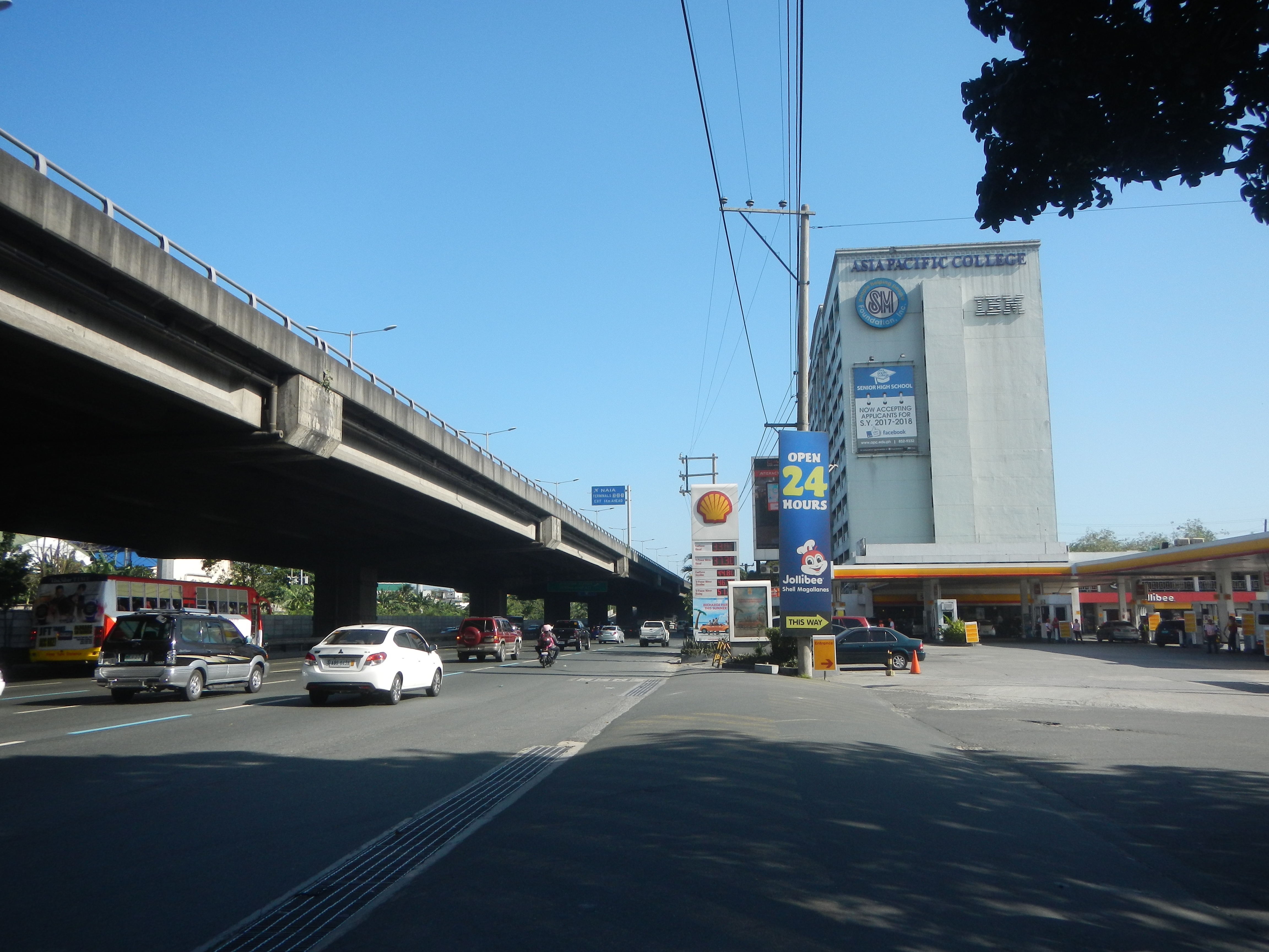 Eine belebte Hauptstraße mit Fahrzeugen, eine Brücke links, Strommasten, Laternen, Bäume, Gebäude mit Namensschildern und eine Tankstelle mit parkenden Autos unter dem Himmel.