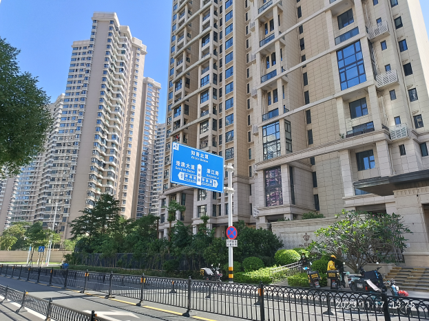 Eine Straße in einer Stadt in Hong Kong mit hohen Wohngebäuden, Bäumen, Pflanzen, einem Zaun, Schildern, Pfosten, Fahrzeugen und einem klaren blauen Himmel.