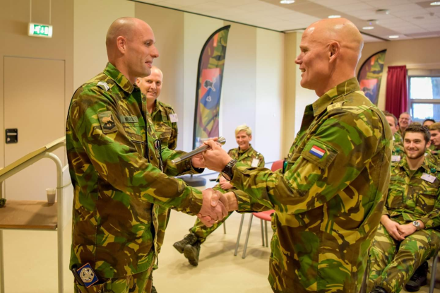 Zwei Männer in Militäruniformen schütteln sich die Hände vor einer sitzenden Gruppe, mit einem Tisch, Geländer, Bannern und einem Fenster mit Vorhang im Hintergrund.