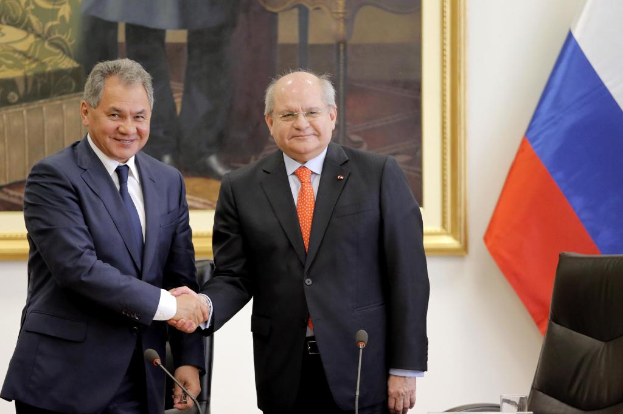 Zwei Männer in Blazern schütteln sich die Hände vor Mikrofonen, mit einem Stuhl, einer Flagge und einem Wandrahmen im Hintergrund.