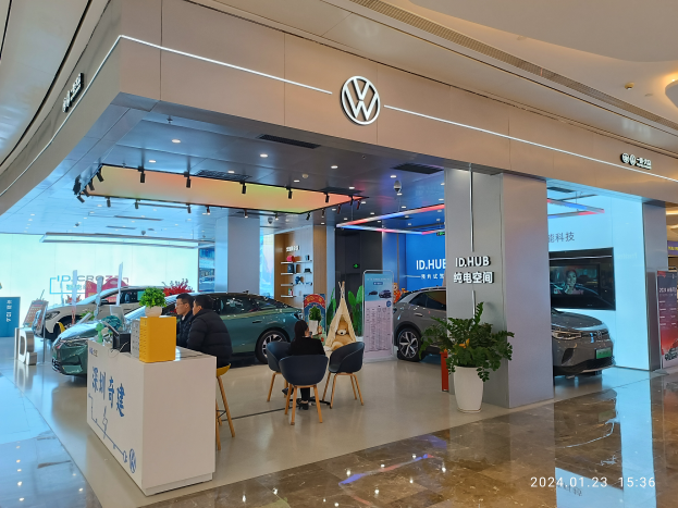 Ein Volkswagen-Händler in Shanghai, China, mit Menschen an einem Tisch mit verschiedenen Gegenständen und Topfpflanzen, Autos und Tafeln mit Text im Hintergrund und einer Decke mit Lampen.