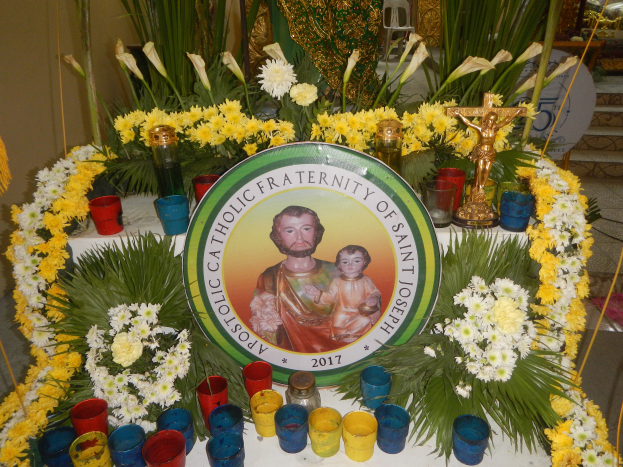 Ein mit bunten Blumen, Kerzen und einer Tafel mit Personalfoto und Text geschmückter Tisch, vor einer Kulisse aus Pflanzen, Stühlen und anderen Gegenständen, schafft eine festliche Atmosphäre für die Bruderschaft des Heiligen Josef.