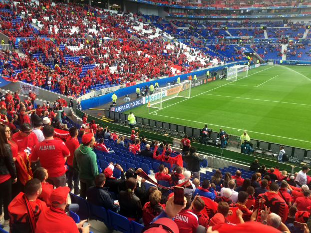Ein volles Stadion mit Zuschauern, einige sitzen und einige stehen, schauen ein Fußballspiel, mit Bannern, einem Torpfosten und Deckenleuchten.
