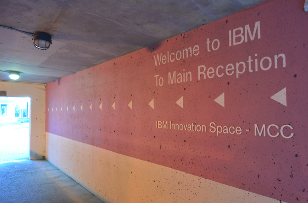 Eine Wand mit den Worten 'Willkommen bei IBM' beleuchtet von Deckenlampen, mit einer Tür zum IBM Innovation Space MCC im Hintergrund.