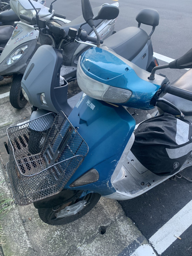 Ein blaues Rollerfahrzeug auf einer Straße in einer Parklöt neben anderen Motorradfahrzeugen geparkt, mit Gras auf der linken Seite.
