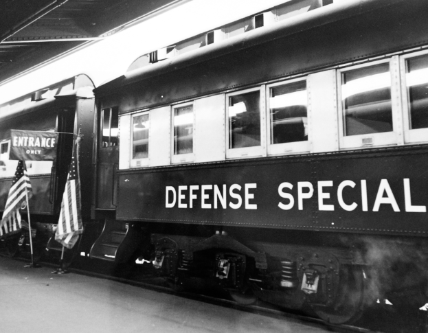 Ein Schwarz-Weiß-Foto eines Zuges mit der Aufschrift 'Defense Special', der auf den Schienen steht und von zwei Fahnen auf der linken Seite und einer Decke darüber umgeben ist.