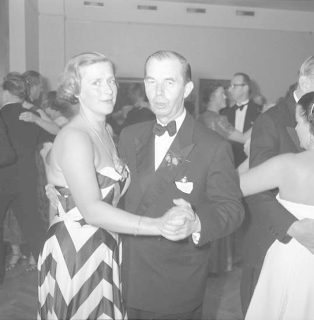 Zwei Menschen, ein Mann in einem schwarzen Blazer, weißem Hemd und schwarzen Hosen und eine Frau in einem unbekannten Outfit tanzen zusammen auf einer formellen Veranstaltung, indem sie sich an den Händen halten.