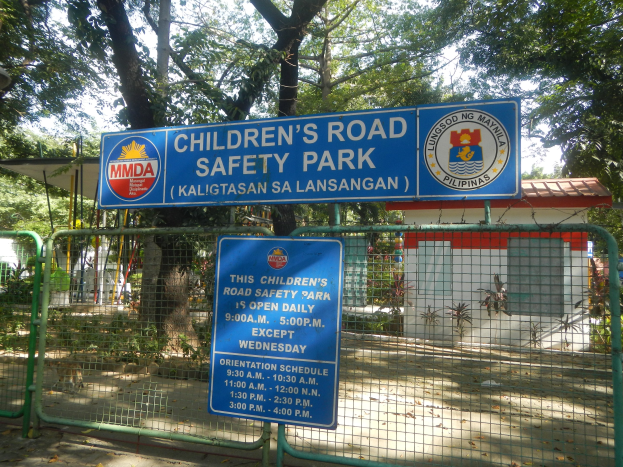 Metallzaun um einen Kinder-Verkehrssicherheitspark mit zwei blauen Informationsschildern, einem kleinen Haus mit Dach, Pflanzen, Bäumen und einem bewölkten Himmel.