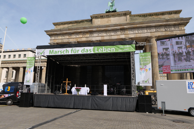 Bühne vor dem Brandenburgischen Tor mit einem Tisch, Bannern, Lautsprechern, Fahrzeugen, Gebäuden, einer Statue, einer Fahne und Wolken.