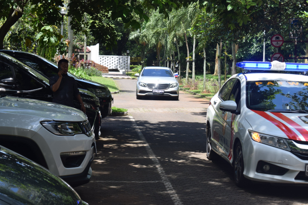 Ein Polizeiwagen steht am Straßenrand neben anderen Fahrzeugen, und eine Person hält in der Nähe ein Mobiltelefon, mit Bäumen, Schildern, Pfählen und einer Wand im Hintergrund.