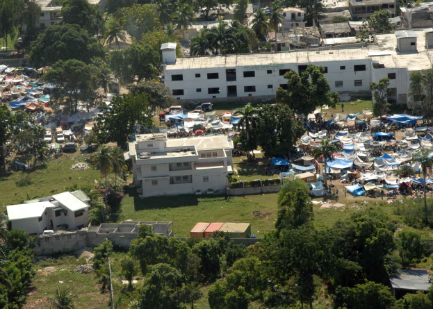 Luftaufnahme einer großen Gruppe von Zelten in einer städtischen Umgebung, wahrscheinlich ein Flüchtlingscamp, umgeben von Gebäuden, Bäumen, Fahrzeugen und anderen städtischen Elementen.