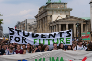 Eine Gruppe von Schülern marschiert in Berlin, die ein buntes "Students for Future"-Schild halten, vor einer Kulisse aus Gebäuden, Bäumen und Himmel.