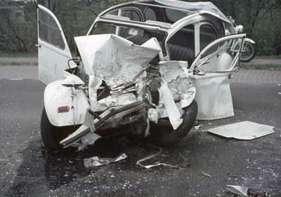 Ein schwerbeschädigtes Auto mit Frontalcrash am Straßenrand, umgeben von Bäumen und einem entfernten Gebäude mit einem weiteren Fahrzeug im Hintergrund.