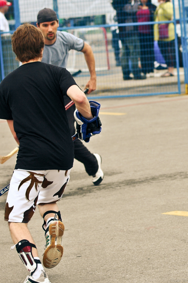 Zwei Hockey spielende Personen auf einer Straße mit einem Zaun dahinter und einer Zuschauermenge im Hintergrund.