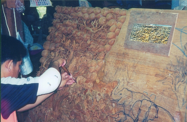 Ein Mann schnitzt an einer Holztafel mit einem Werkzeug und stellt dabei Elefanten und Bäume dar, im Hintergrund sind ein Kalender, eine Tasche und ein Holzstab zu sehen.