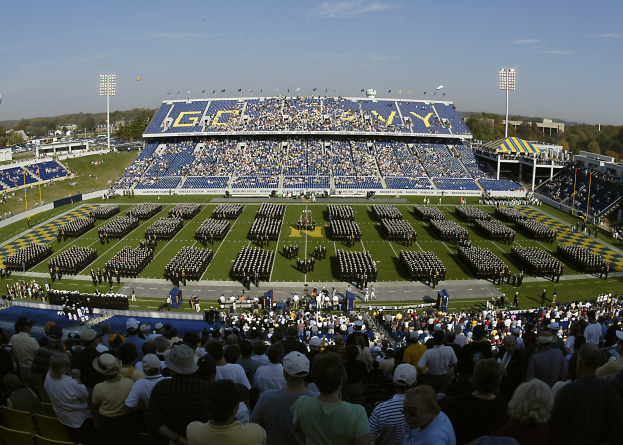 Eine große Menschenmenge in einem Stadion, die ein Fußballspiel verfolgt, wobei einige stehen und andere sitzen, auf einem Rasenfeld unter einem klaren blauen Himmel, mit Flutlichtmasten, Bäumen und Gebäuden im Hintergrund.