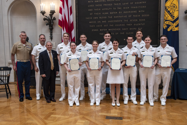 Gruppe von Menschen in Uniform, die strahlen und Zertifikate halten, mit einem Tisch, der mit blauer Stoff bedeckt ist, Stühlen, Fahnen, Lichtern, einer Tafel mit der Aufschrift "U.S. Naval Academy Class of 2018" und einer Wand im Hintergrund.