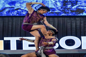 Zwei Akrobaten führen auf der Bühne eine Vorstellung auf, wobei der Mann die Frau in lila Kleidern hochhebt, während das Publikum unten zuschaut.