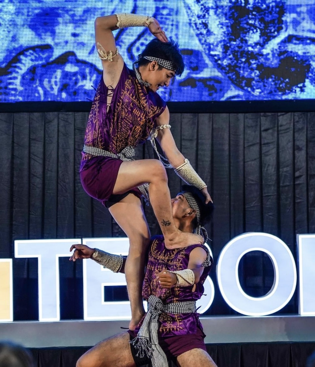 Zwei Akrobaten führen auf der Bühne eine Vorstellung auf, wobei der Mann die Frau in lila Kleidern hochhebt, während das Publikum unten zuschaut.