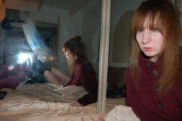 Eine Frau in roter Jacke steht neben einem Spiegel, der ihr Bild reflektiert, mit einem Bett und Laken im Hintergrund.