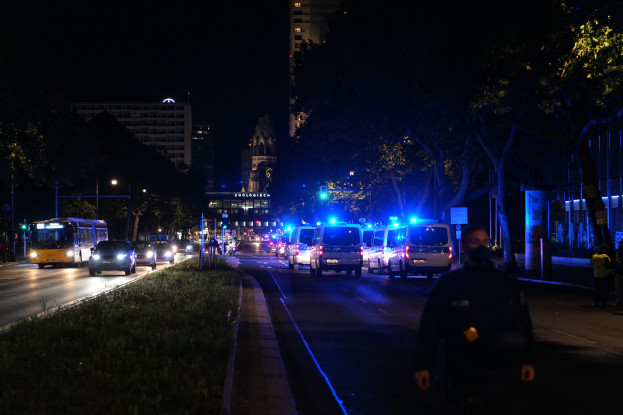 Eine nächtliche Straßen Szenerie mit Polizeiwagen und Bussen, Menschen auf dem Bürgersteig, Gras auf der linken Seite, Bäume und Gebäude im Hintergrund und Laternen, die die Straße beleuchten.
