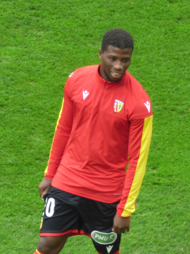 Ein Mann in einem roten T-Shirt und schwarzen Shorts steht auf einem Fußballfeld, lächelt und ist bereit, als Flügelspieler zu spielen.