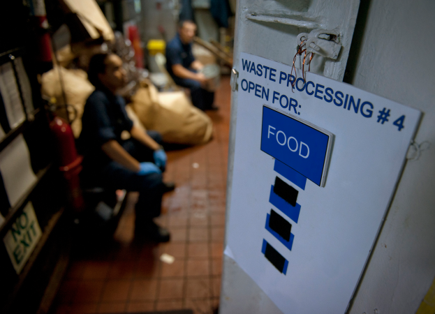 Ein Mann sitzt auf dem Boden neben einem Schild mit der Aufschrift "Abfallverarbeitung 4: Open for Food", mit einer Tür und einem Papier auf der rechten Seite, Regalen mit Gegenständen auf der linken Seite und unscharfen Figuren und Taschen im Hintergrund.