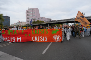 Eine Gruppe von Menschen marschiert mit einem Banner, auf dem "Klima-Krise ist eine Krise" steht, eine Straße entlang, mit Bäumen und Gebäuden gesäumt und einer Brücke im Hintergrund unter einem bewölkten Himmel.
