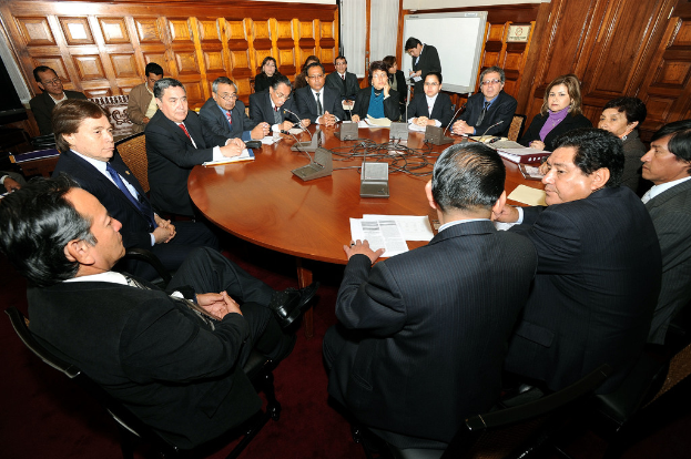 Eine Runde Tischbesprechung mit Delegierten, die über Mikrofone, Papiere, Kabel und Dateien auf dem Tisch sprechen, mit einer hölzernen Wand im Hintergrund.