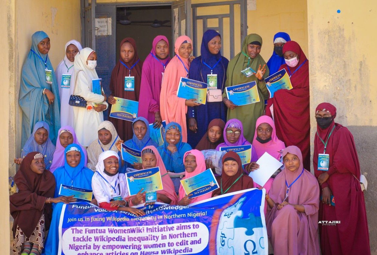 Eine Gruppe von Frauen, die zusammenstehen, lächeln und Zertifikate halten, vor einem Gebäude mit einem Banner, auf dem "Funtua Women Wiki Initiative möchte die Wikipedia-Ungleichheit im nördlichen Nigeria angehen".