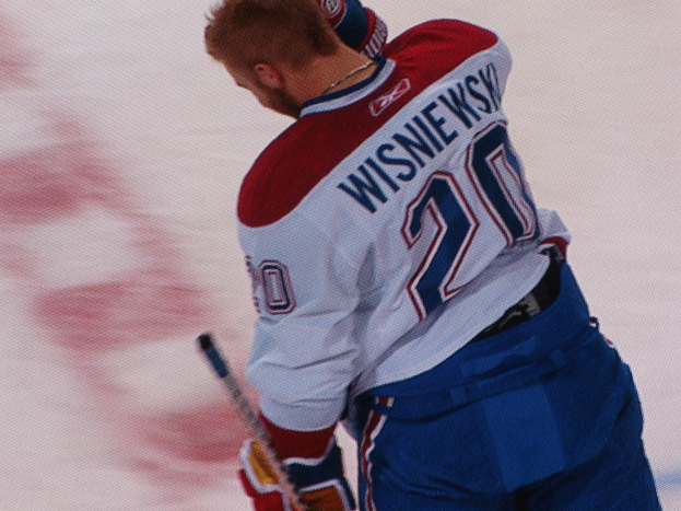 Ein Hockey-Spieler in einem "Wisniewski"-Trikot steht auf dem Eis, hält einen Hockey-Schläger und hat einen konzentrierten und entschlossenen Gesichtsausdruck.