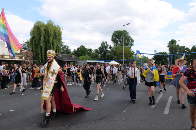 Eine Gruppe von Menschen marschiert auf einer Straße während der Pride Parade 2018, einige tragen eine Regenbogenflagge und Musikinstrumente, mit Laternenmasten, Bäumen, Hütten und einem bewölkten Himmel im Hintergrund.