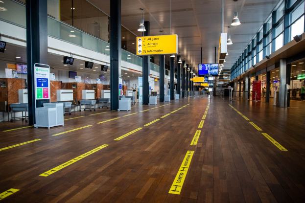 Langer Flughafenflur mit gelbem Absperrband auf dem Boden, Säulen, Informationsschilder, Bildschirme, Lampen, Glaswände und eine Person im Hintergrund.