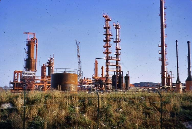 Ölraffinerie in einer grünen Wiese mit Hügeln und einem klaren Himmel, umgeben von Zäunen, Pfählen, Tanks und anderen industriellen Strukturen.