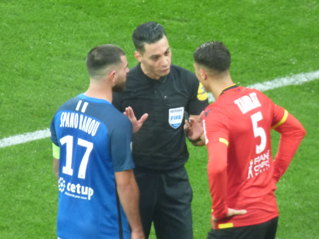 Ein Schiedsrichter in schwarzer Kleidung spricht mit zwei Männern auf einem Fußballfeld, einer in blau und rot Hemden mit Text, beide tragen Brillen.