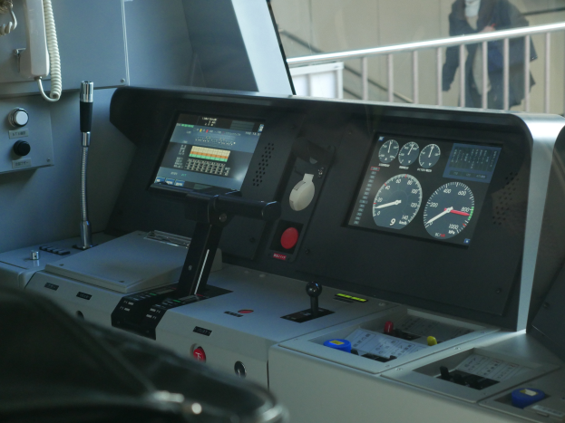 Ein Nahaufnahme eines Zugsteuerpults mit Knöpfen und Drehreglern, ein Telefon auf der linken Seite und eine Person, die in der Nähe eines Geländers steht.